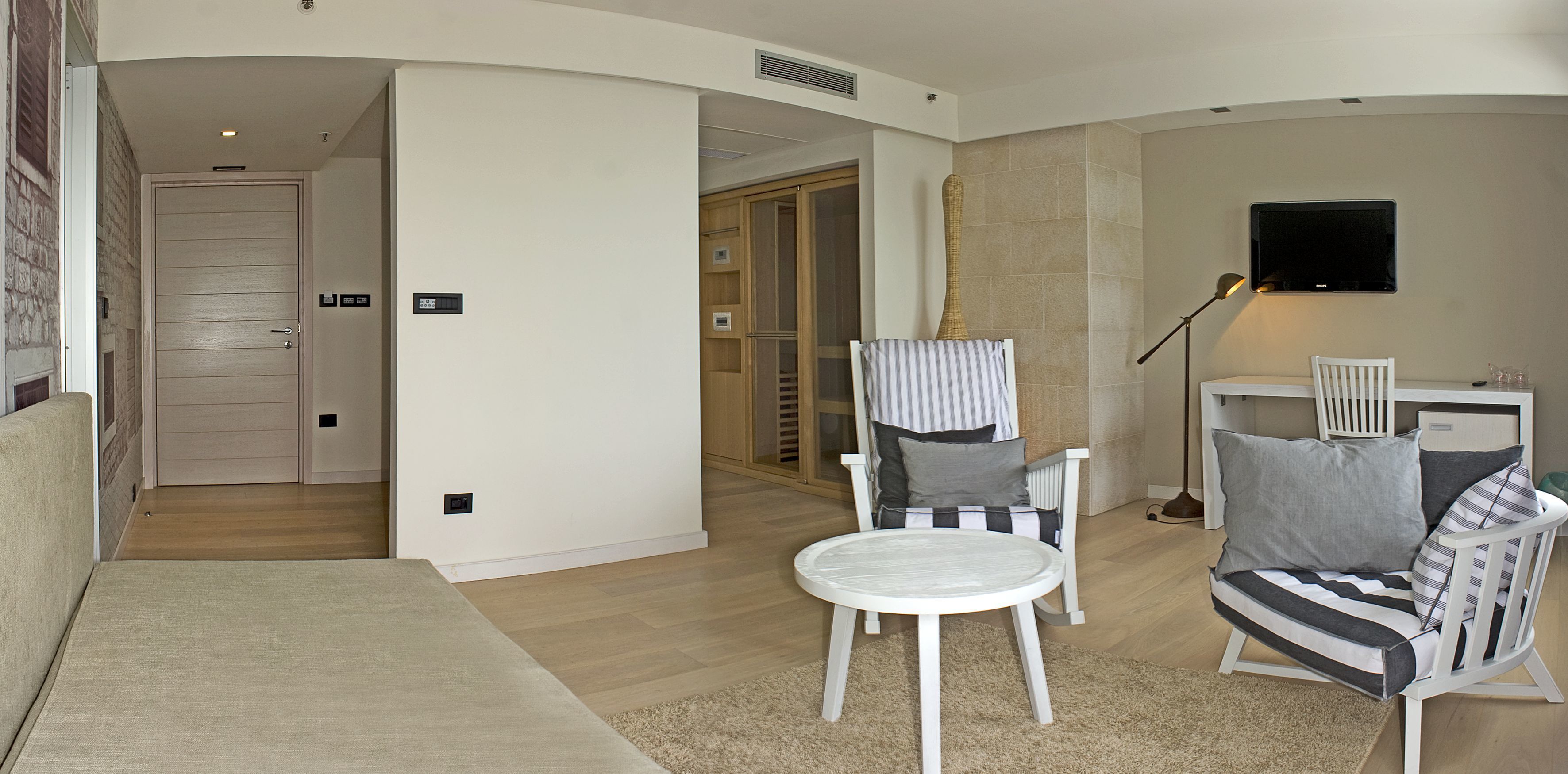 Un salon moderne avec un canapé blanc, un fauteuil à rayures et une table ronde. Un téléviseur est fixé au mur et une lampe se trouve sur une table d’appoint.