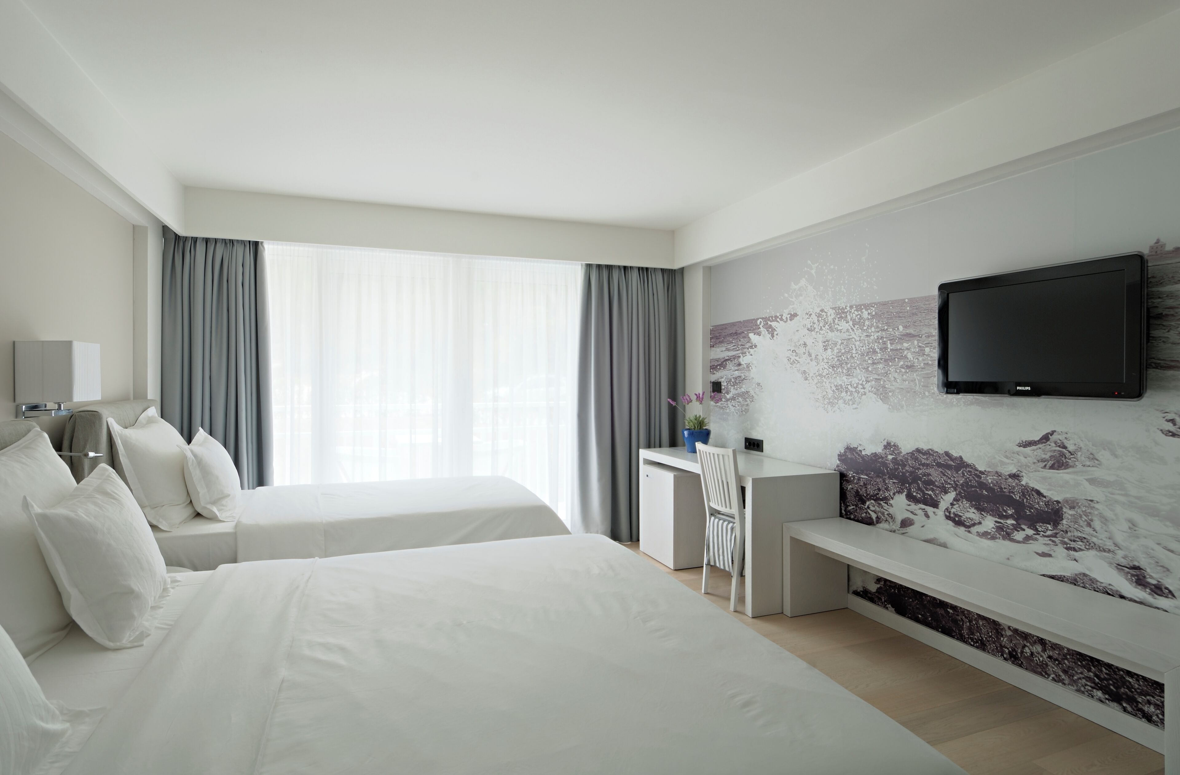 Ein Hotelzimmer mit zwei Betten, einem Schreibtisch, einem Stuhl, einem Fernseher und einem großen Fenster mit Vorhängen.
