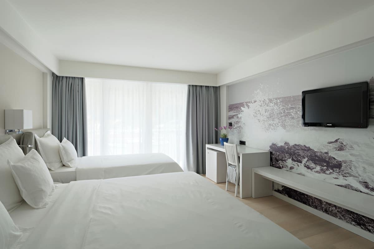 A hotel room with two beds, a desk, a chair, a TV, and a large window with curtains.