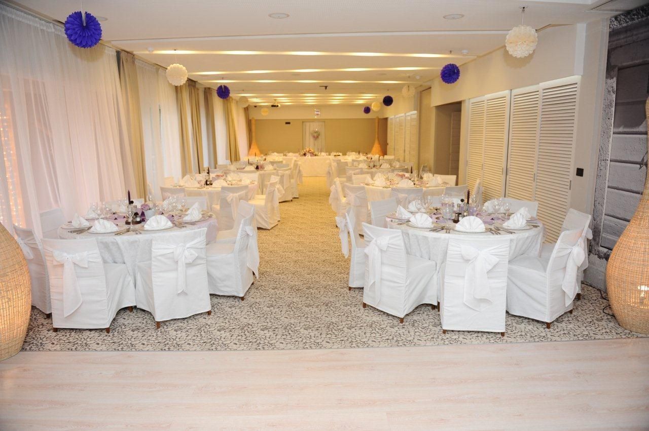 Een balzaal met ronde tafels gedekt voor een formeel evenement, met witte tafelkleden en stoelen, en een neutraal kleurenschema.