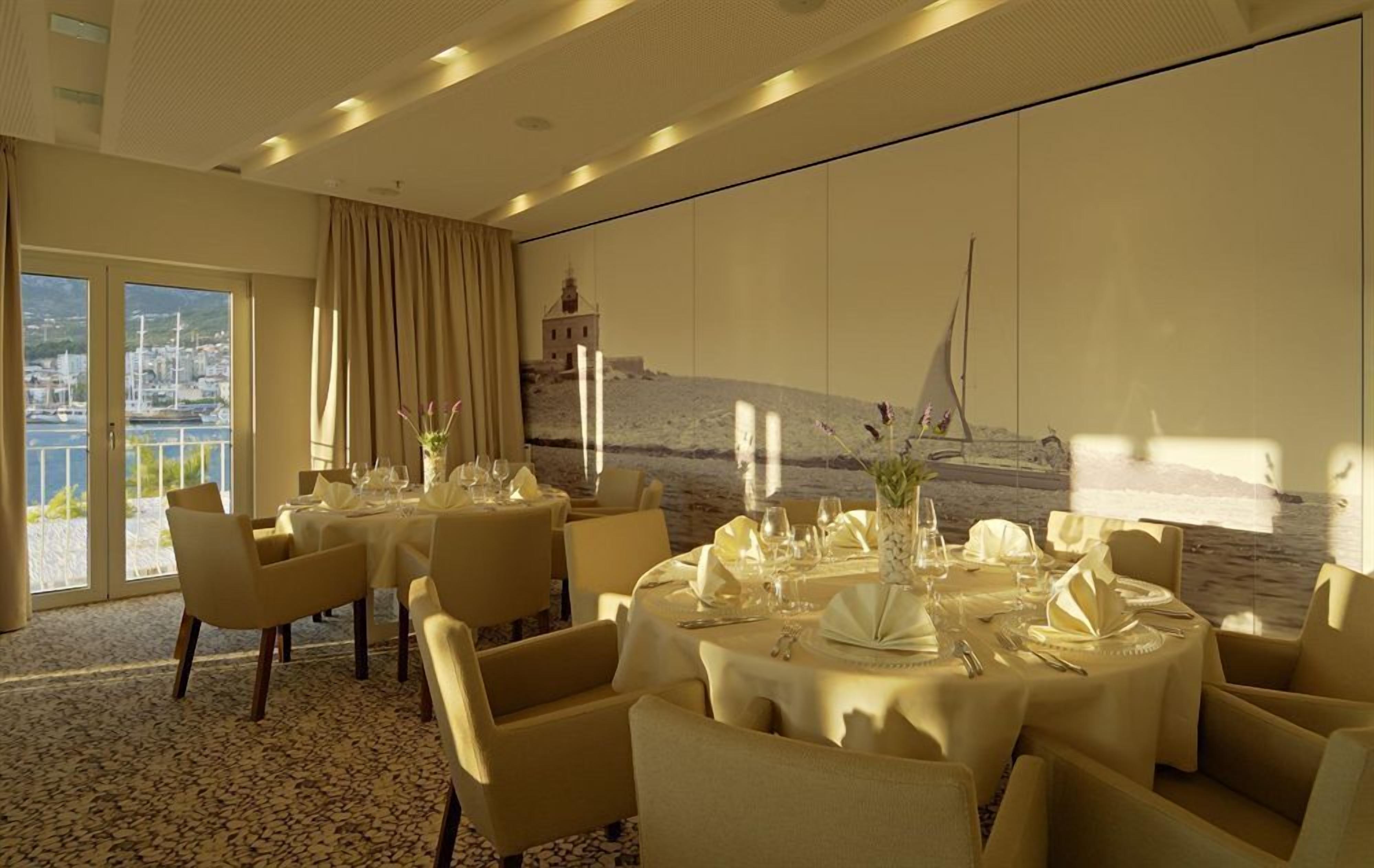Une salle à manger avec des tables rondes disposées pour accueillir quatre personnes, chacune étant recouverte d’une nappe blanche et agrémentée de verres et de couverts. Des chaises beiges sont placées autour des tables. Une grande fenêtre offre une vue sur un port et sur des bâtiments.