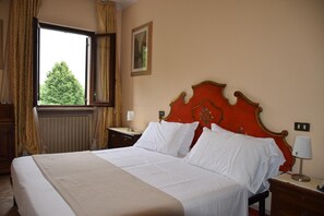 Een hotelkamer met een groot bed, een nachtkastje, een raam met gordijnen, een radiator en een lijst met een afbeelding aan de muur.