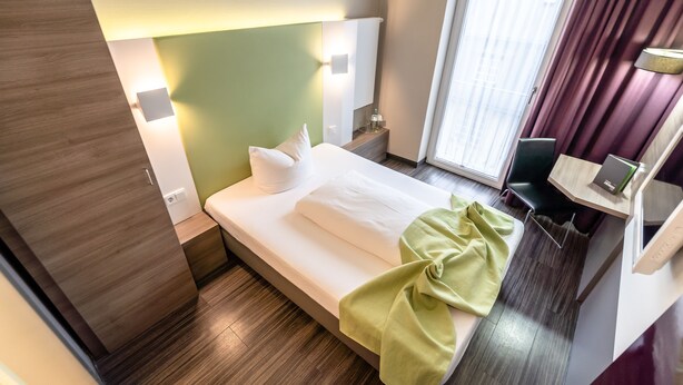 Habitación de hotel con cama, mesita de noche, lámpara, silla y ventana con cortinas.