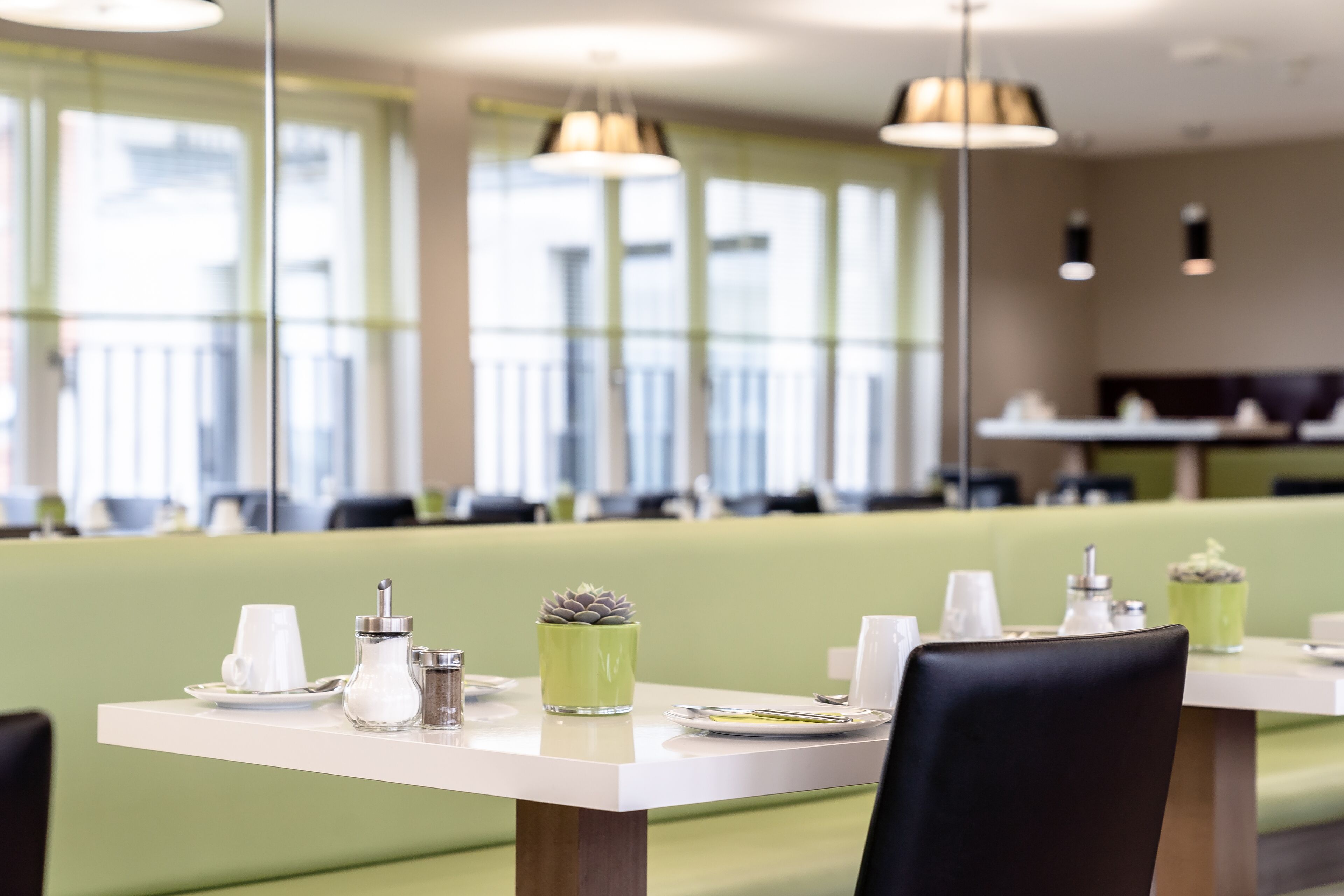 Un restaurant avec des tables dressées pour le service, comprenant un coin salon avec des banquettes vertes et des lampes suspendues.