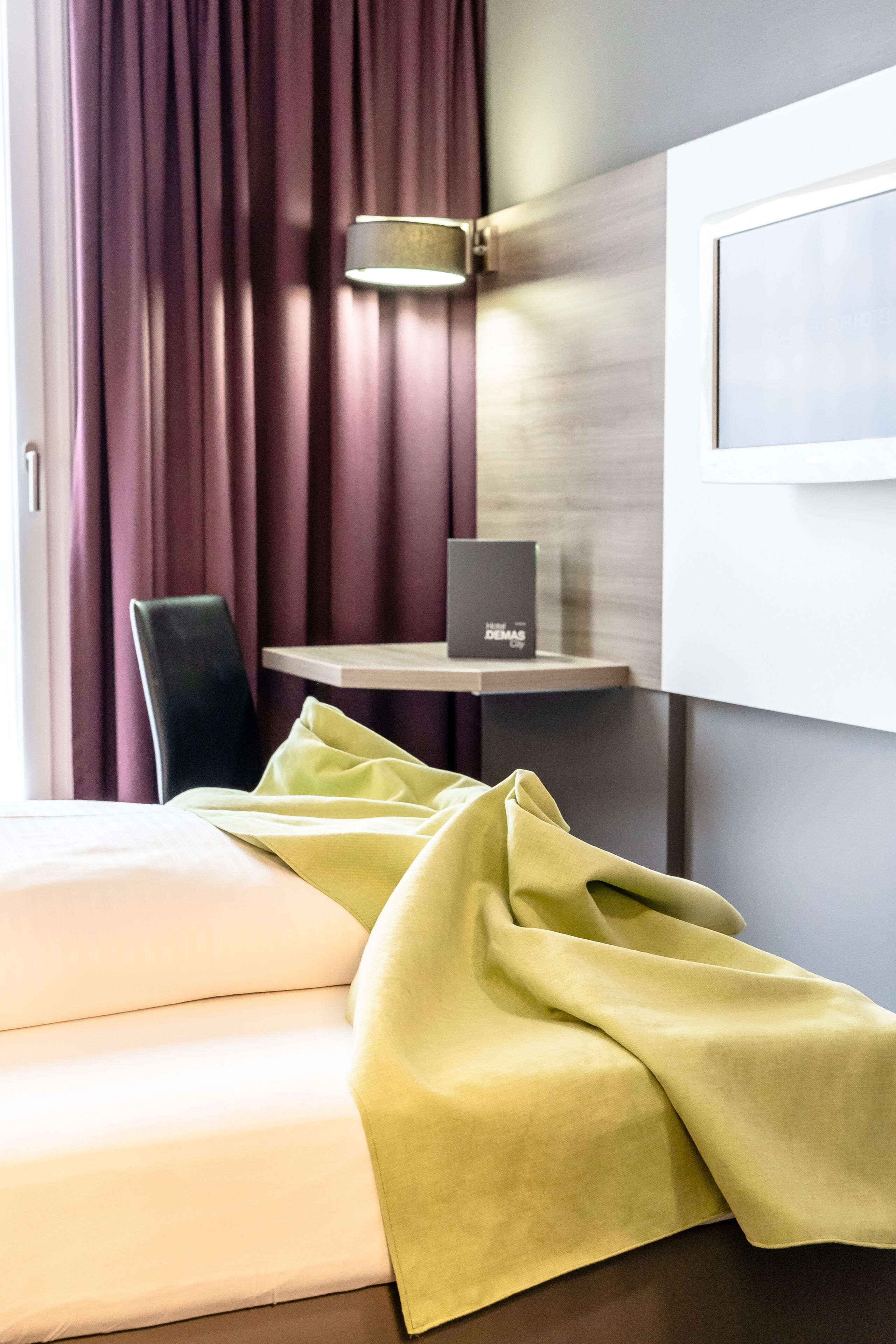 A hotel room with a bed, a chair, a table with a lamp, and a wall-mounted screen.