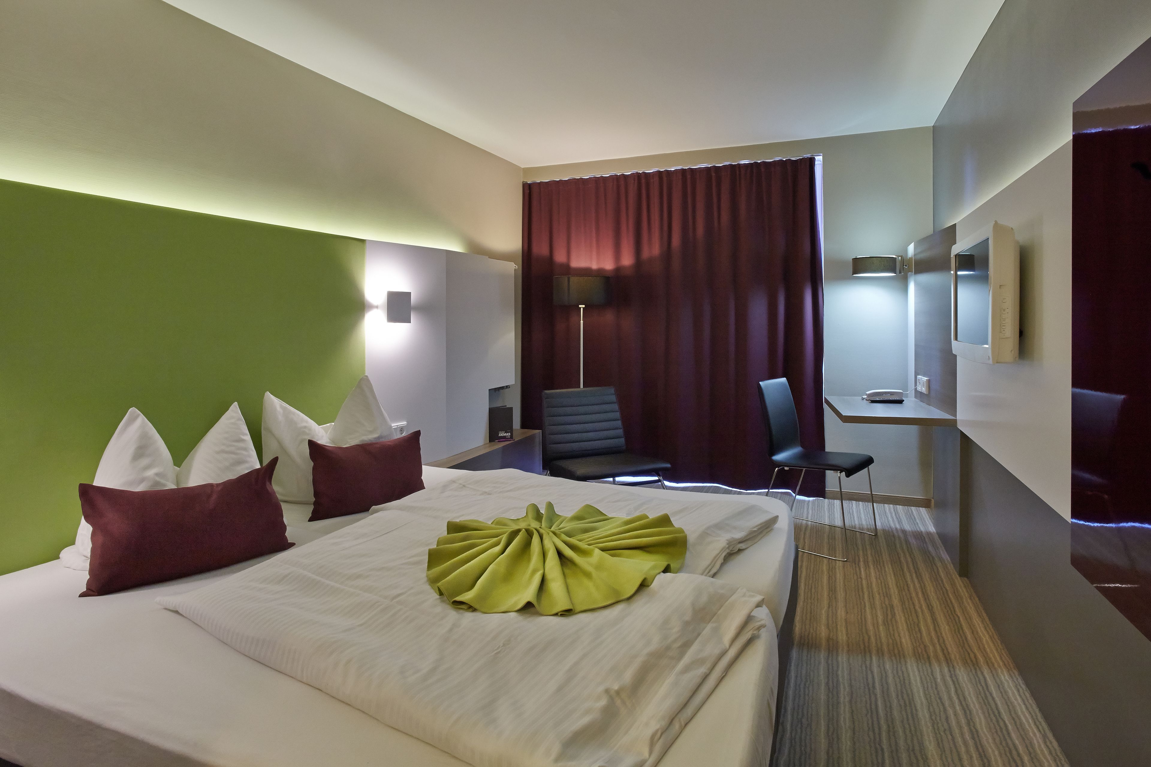 A hotel room with a neatly made bed, a desk with a chair, a TV, and a lamp.
