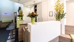 Un área de recepción moderna con un mostrador blanco, dos jarrones con flores verdes y una zona de bar con taburetes negros.