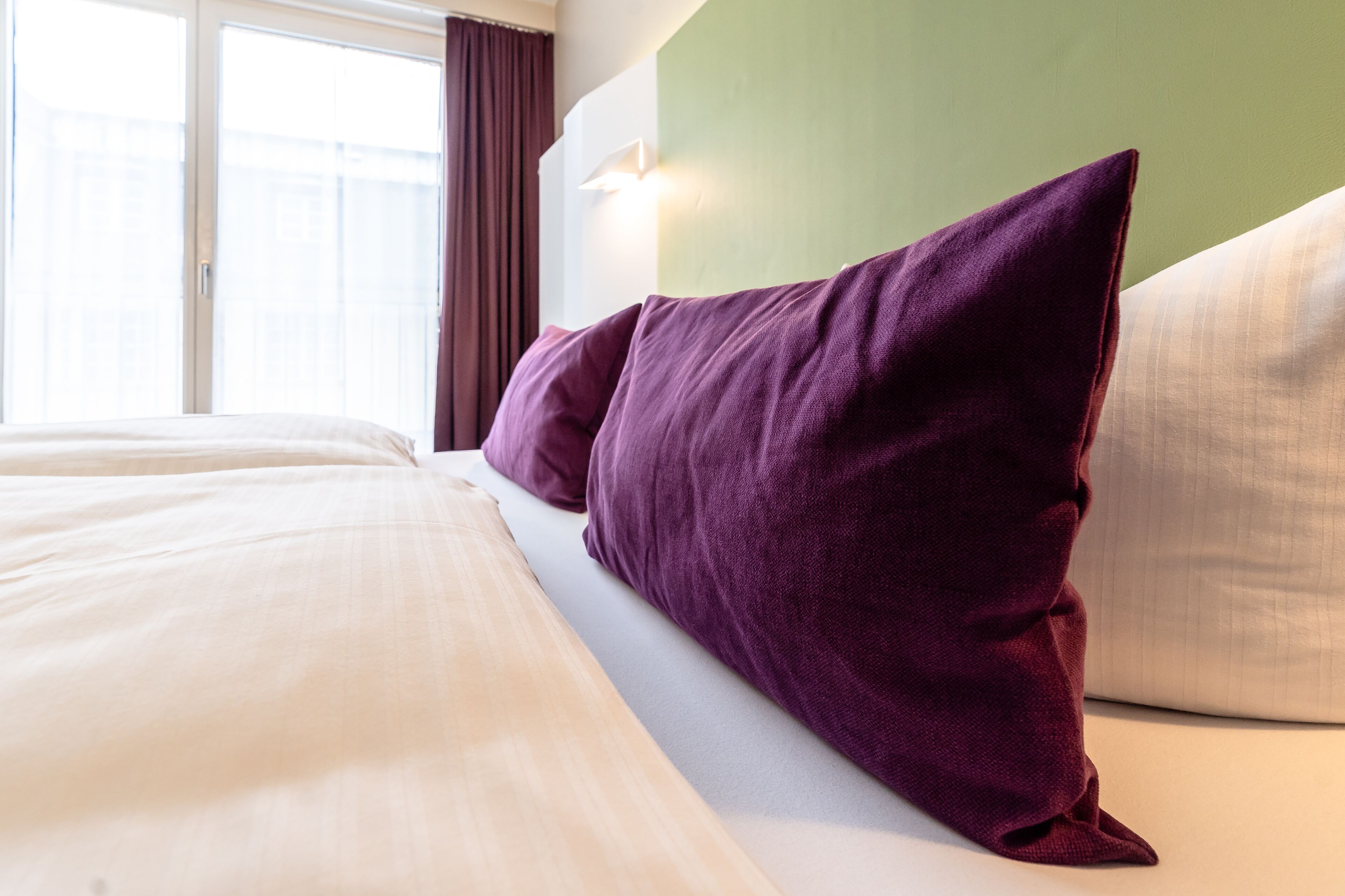 A hotel room with a bed, purple and white pillows, a green wall, and a window with curtains.