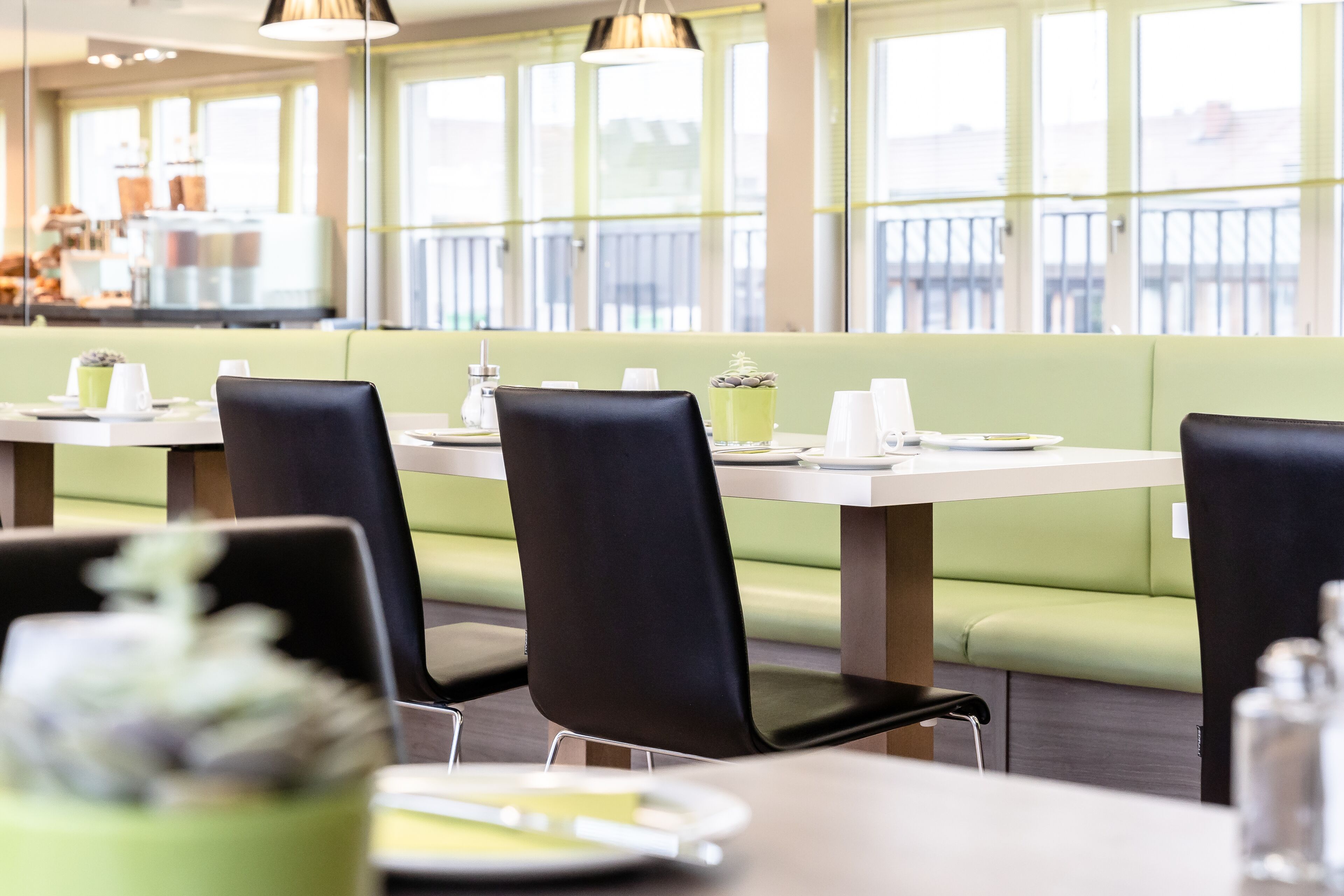 A restaurant with green booths and black chairs, white tables, and a view of the city through large windows.