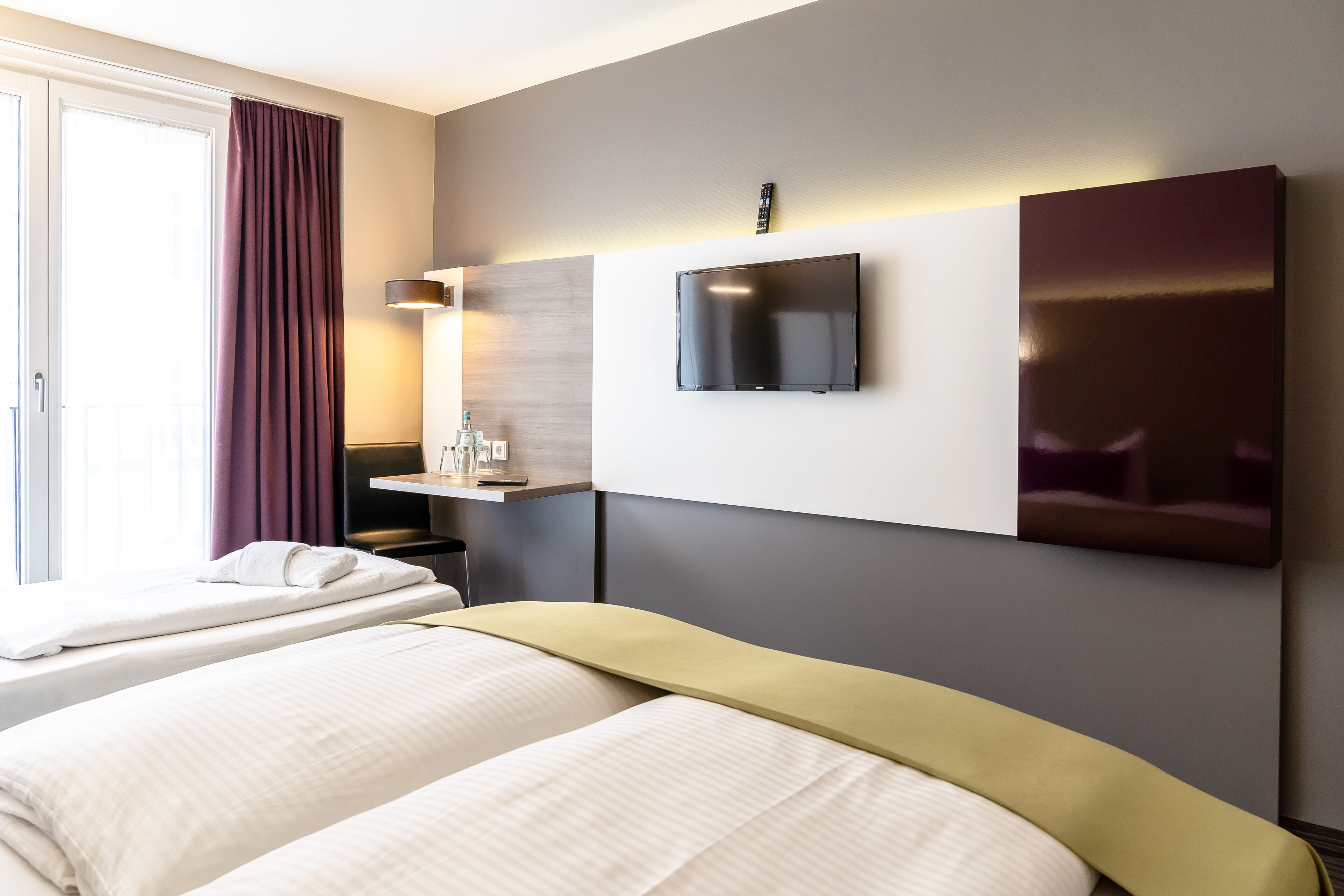 A hotel room with two beds, a TV mounted on the wall, a desk with a lamp, and a window with curtains.