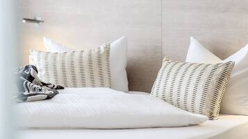 Una cama bien hecha con ropa blanca y almohadas a rayas, una silla de mimbre y una pared de color claro.
