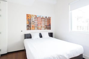 Quarto de hotel com cama, janela e pintura de paisagem urbana na parede.