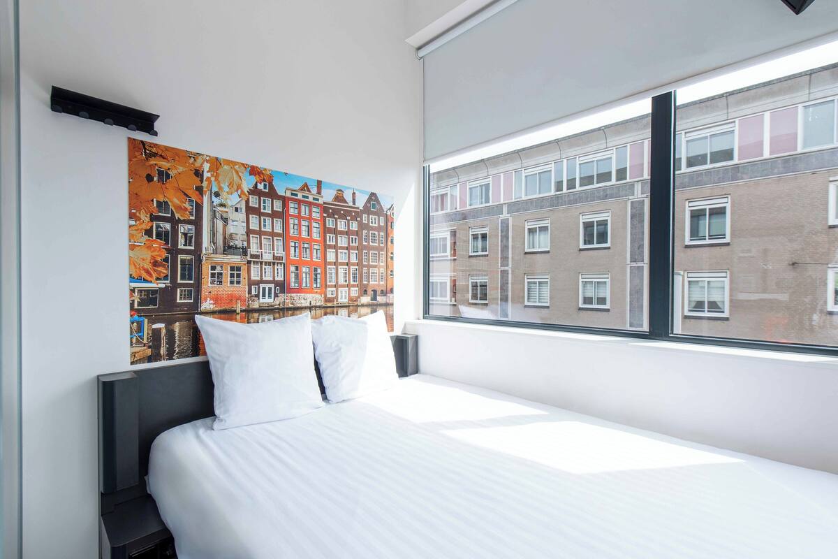 Habitación de hotel con un ventanal grande, cama con ropa blanca y un mural de un paisaje urbano colorido en la pared.