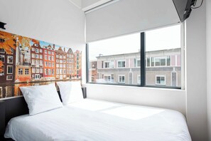 Une chambre d’hôtel avec un grand lit, du linge de lit blanc, une fenêtre donnant sur les bâtiments de la ville et une fresque colorée sur le mur.