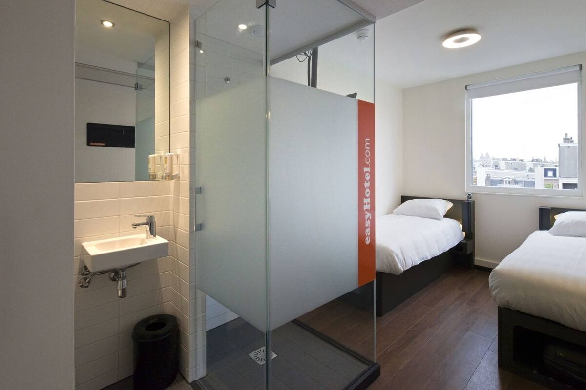 Habitación de hotel moderna con separador de vidrio, lavamanos, cama y ventana con vista a la ciudad.