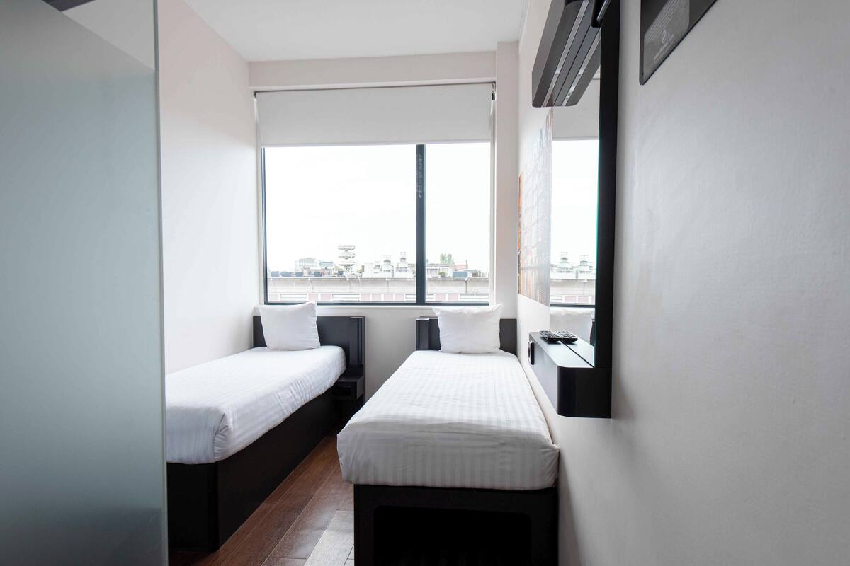 Una habitación de hotel pequeña con dos camas individuales, una ventana con vista a la ciudad, un escritorio con silla y un televisor montado en la pared.