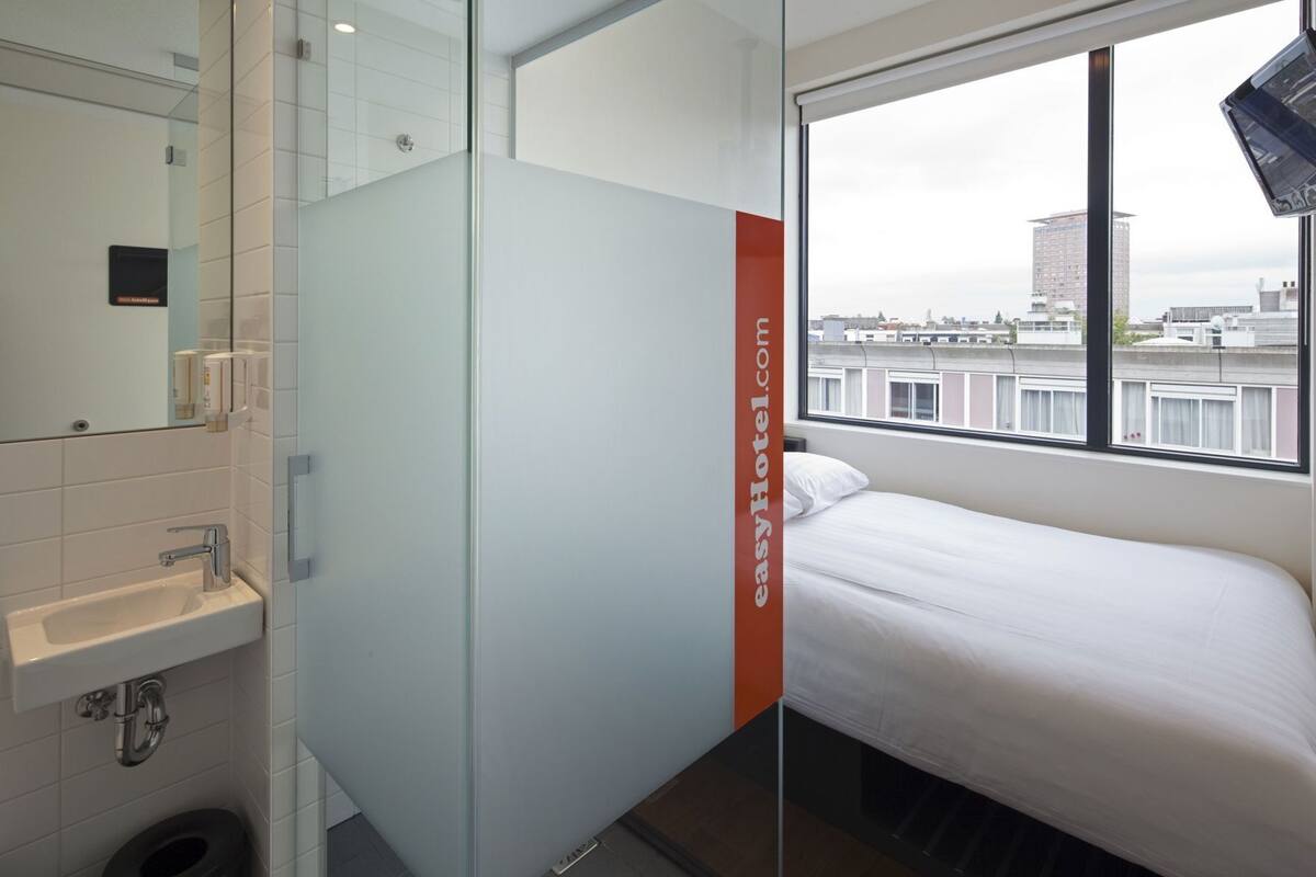 Una habitación de hotel con cama, ventana con vista a la ciudad, lavamanos y espejo.