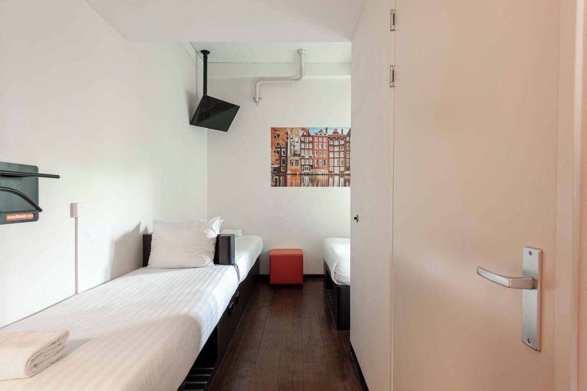 Habitación de hotel con dos camas, un reposapiés rojo, un televisor montado en la pared y un cuadro de edificios.