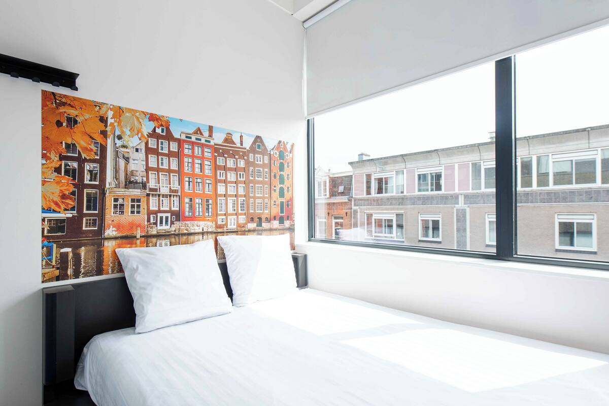 Habitación de hotel con un ventanal grande, una cama con ropa de cama blanca y un mural de un paisaje urbano colorido en la pared.