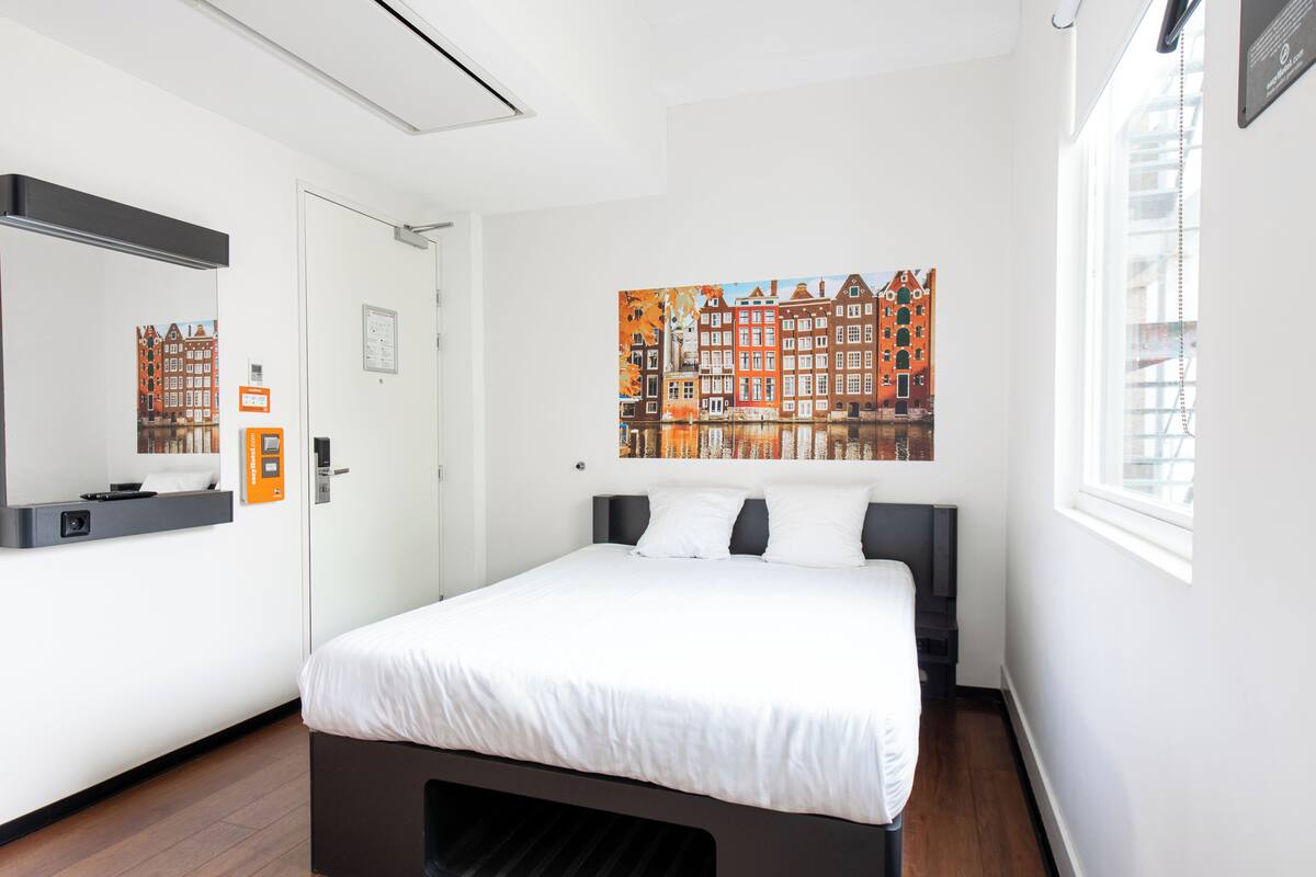 Una habitación de hotel moderna con una cama grande, un espejo, un cuadro de un paisaje urbano y una ventana con persianas.