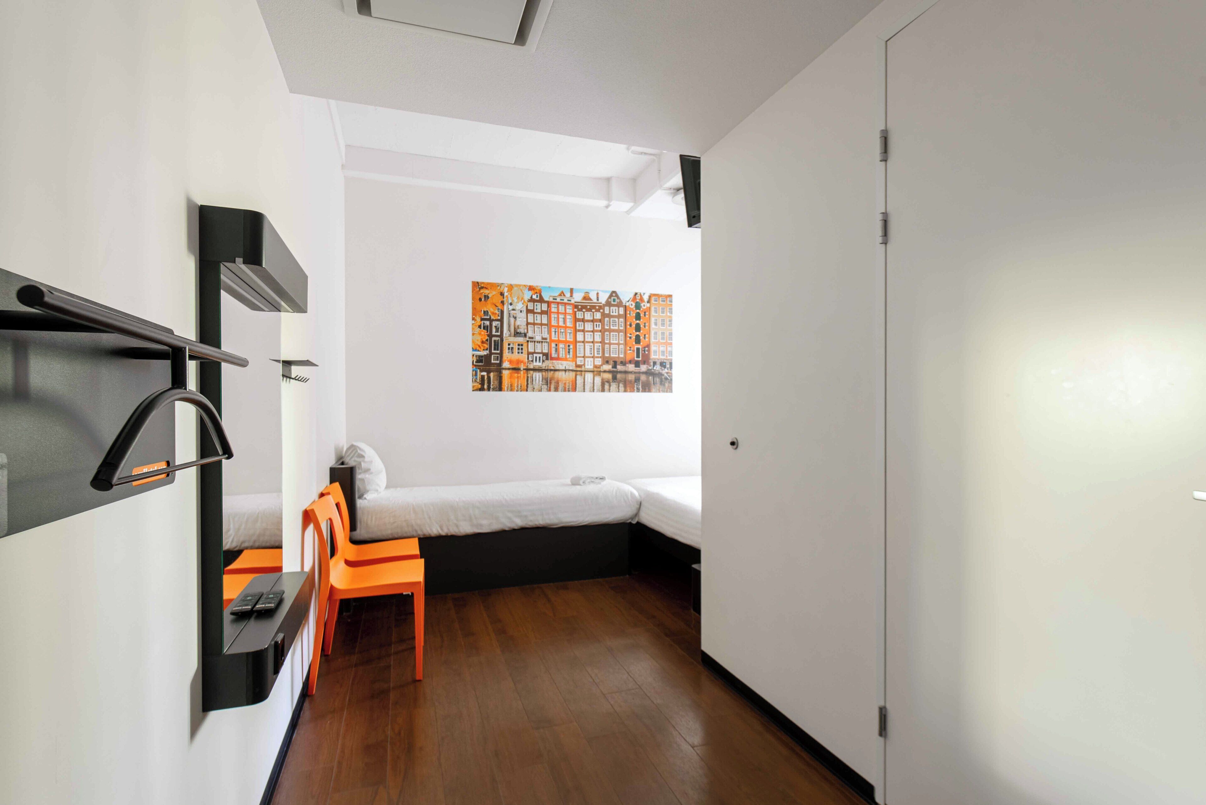 A compact room with a bed, a small table, a chair, a wall-mounted shelf, and a painting of a cityscape.