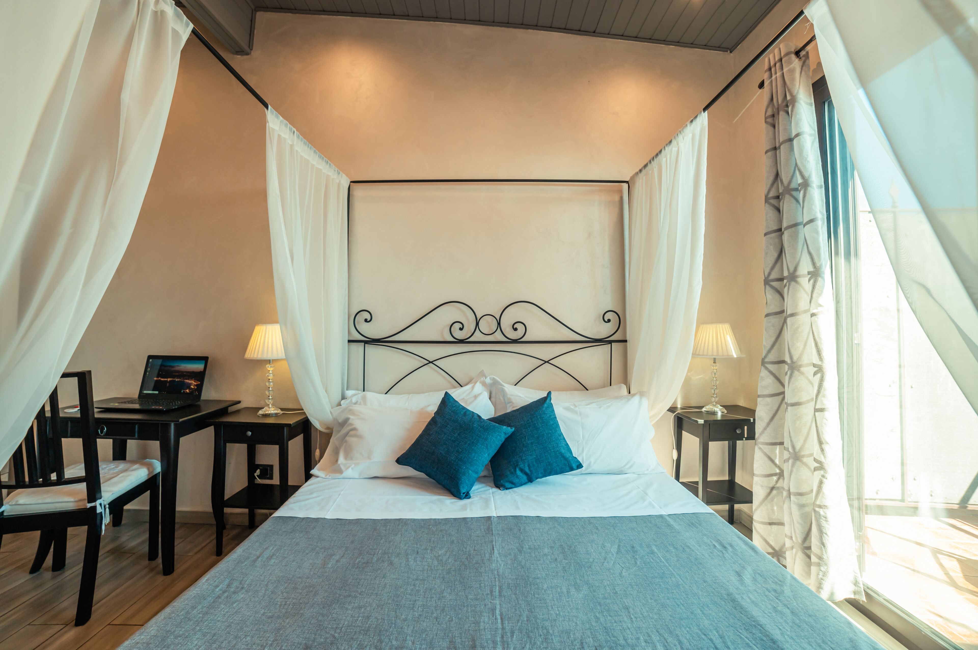 A bedroom with a four-poster bed, two bedside tables with lamps, a laptop on one table, and a chair.