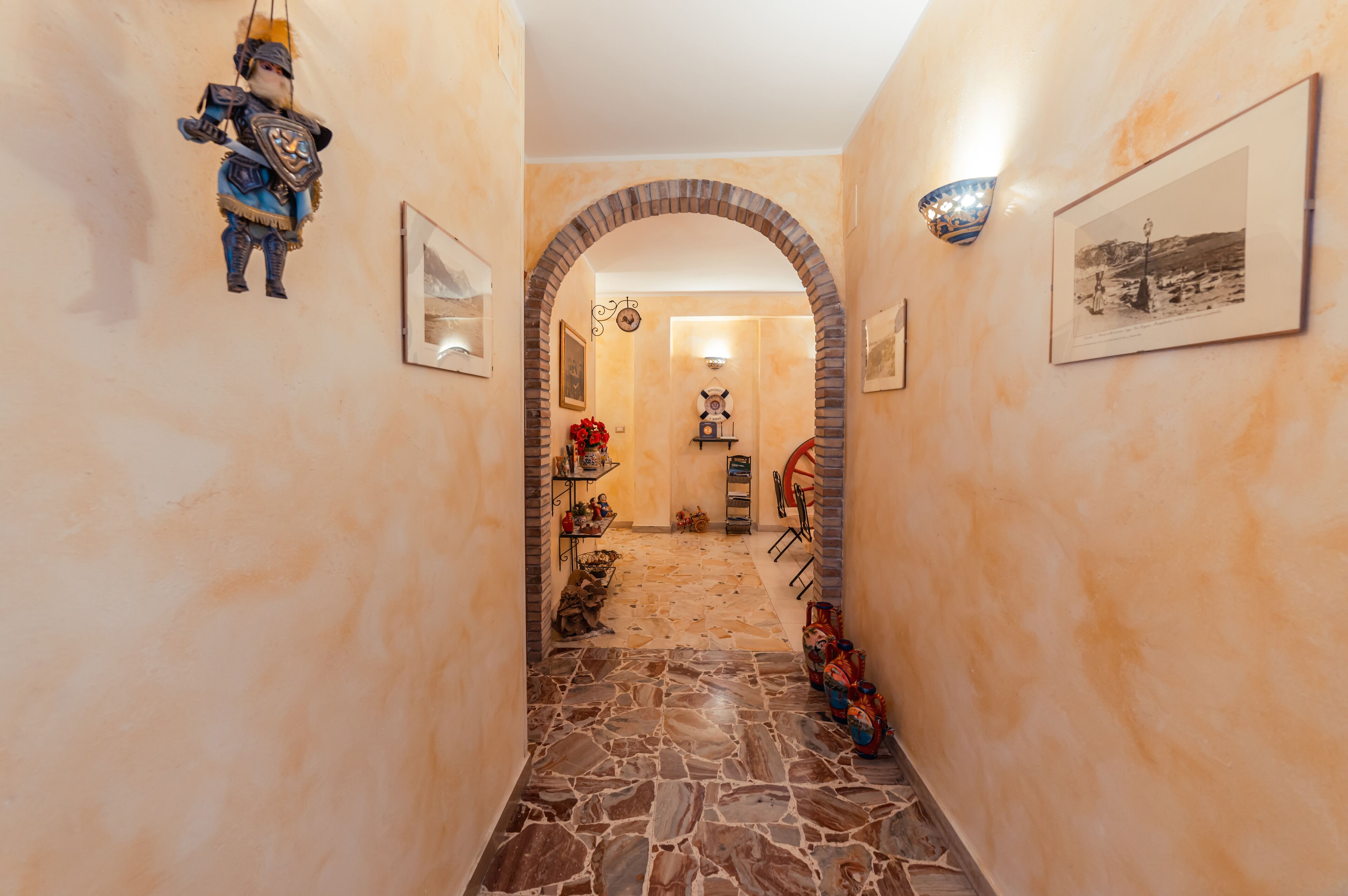 A hallway with stone flooring, a brick archway, and various decorations including a framed picture, a wall-mounted light fixture, and a hanging ornament.