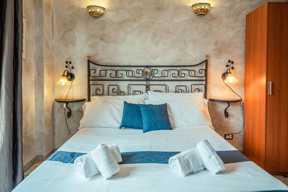 A neatly made bed with white and blue bedding, two wall-mounted lamps, and a wooden wardrobe.