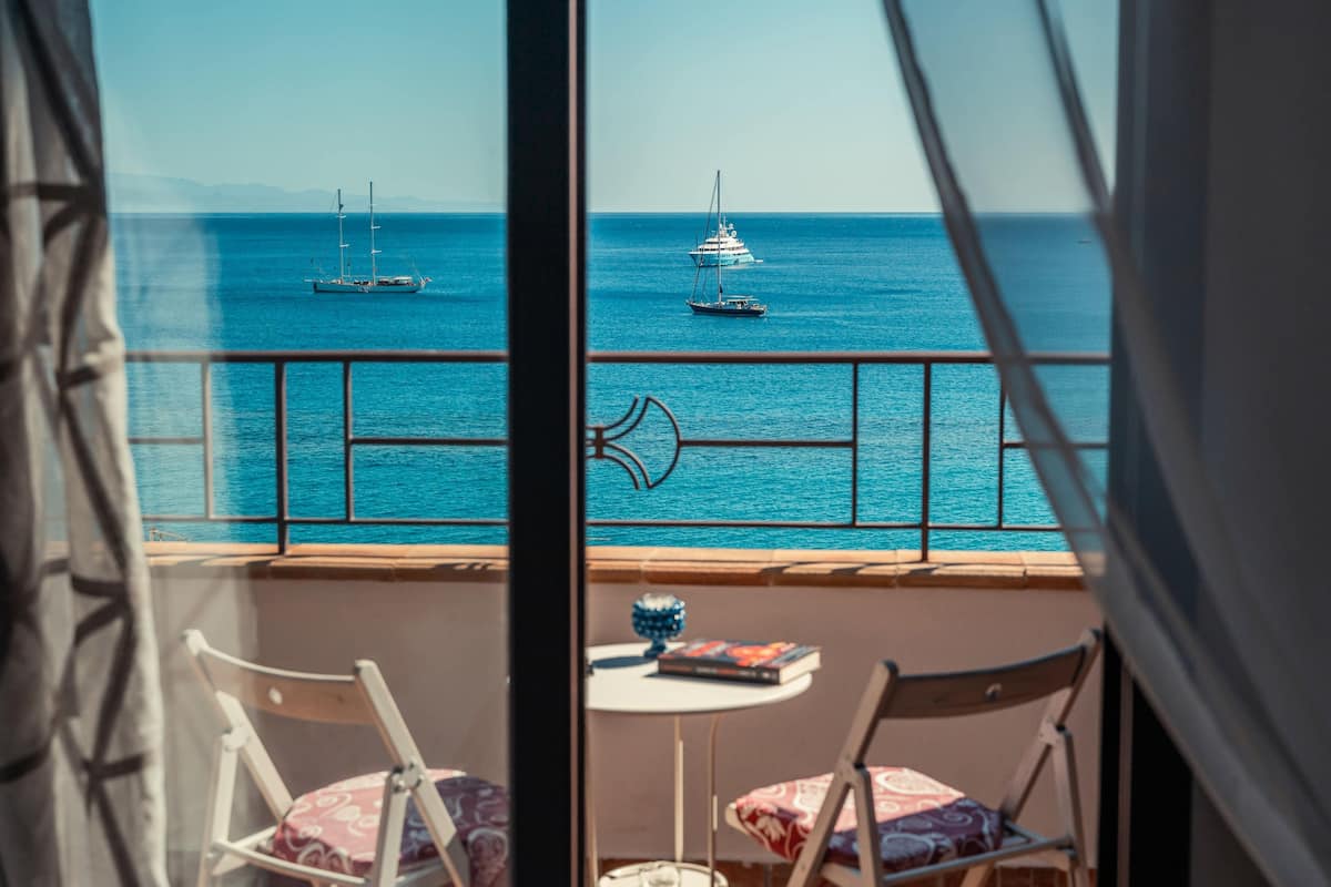 A balcony with two chairs and a small table, overlooking a calm sea with sailboats.