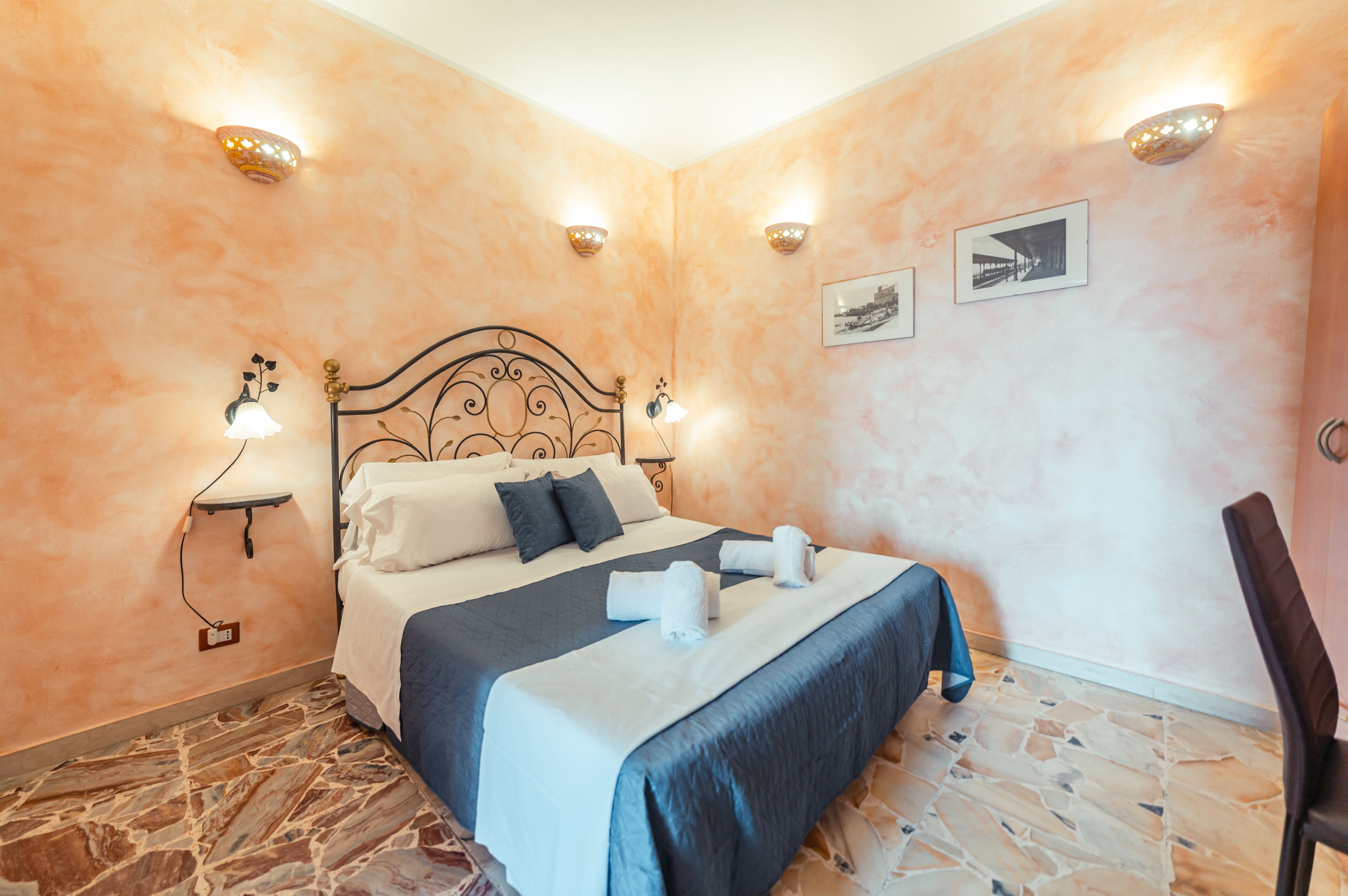 A neatly made bed with a headboard, two wall-mounted lights, and a small shelf with a lamp.