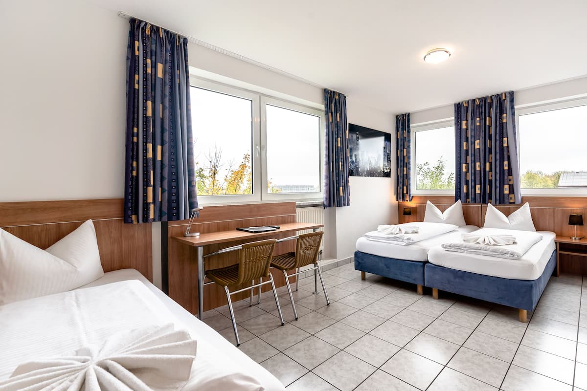 A hotel room with a bed, a desk, two chairs, a TV, and large windows with curtains.