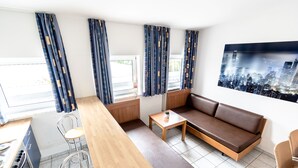 A modern living room with a sofa, chairs, a dining table, and a kitchen area. - Hotel Fresh INN (Unterhaching)