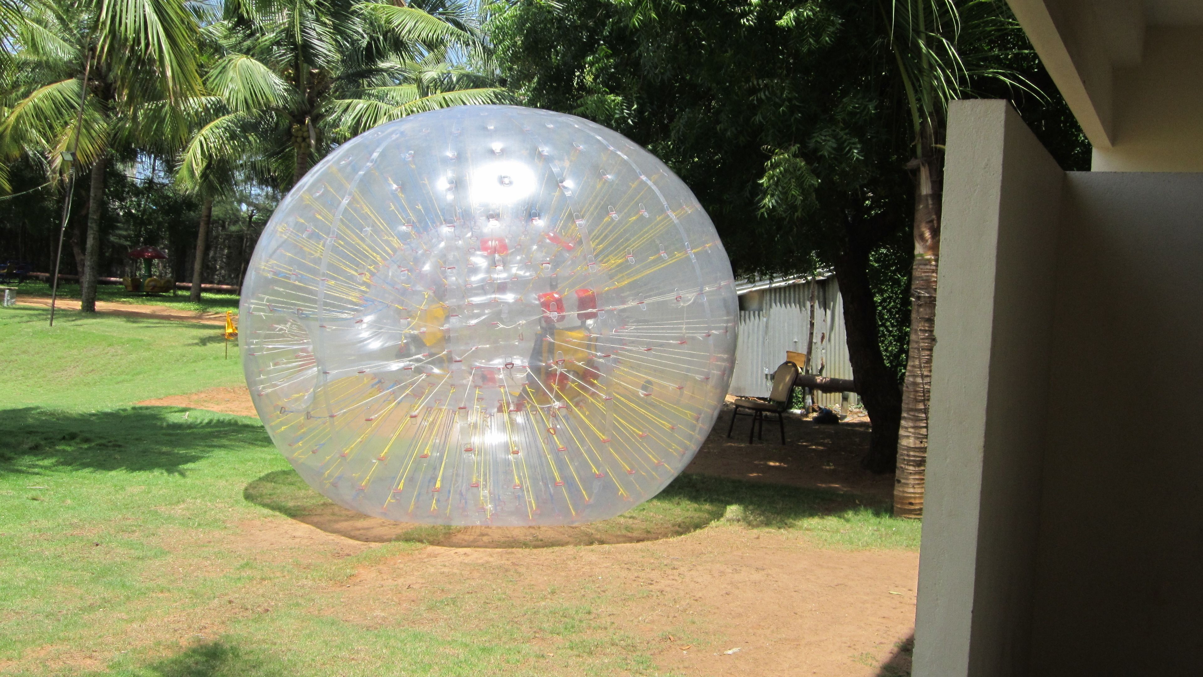 A large transparent ball with yellow poles and red clips, situated outdoors on a grassy area with trees and a building in the background.
