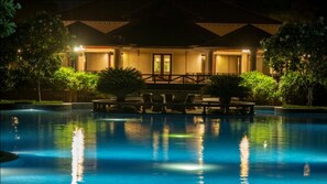 A nighttime view of a hotel with a swimming pool, outdoor seating, and a well-lit entrance.