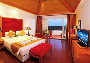 A hotel room with a large bed, a television, a balcony with curtains, and a wooden floor.