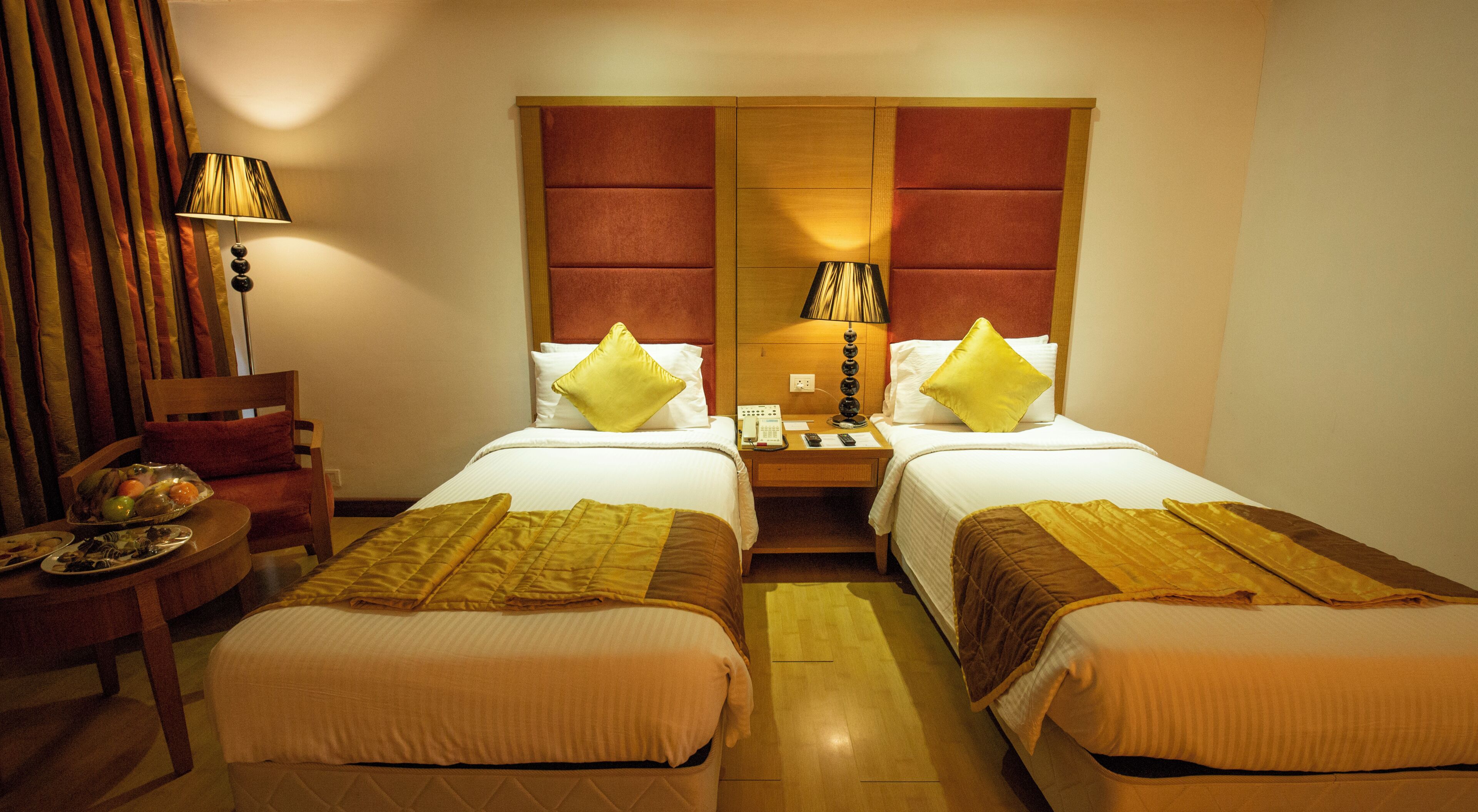 Habitación de hotel con dos camas, cabecera de madera, mesita de noche con lámpara, una silla y una pequeña mesa con fruteros.