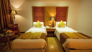 Habitación de hotel con dos camas, cabecera de madera, mesita de noche con lámpara, una silla y una pequeña mesa con fruteros.