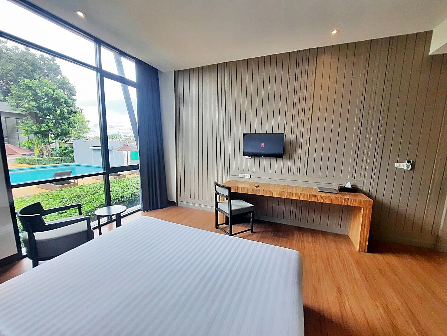 Habitación de hotel moderna con una cama grande, un escritorio de madera, una silla, un televisor montado en la pared y vista a una piscina exterior.