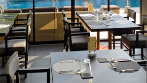 A dining area with tables and chairs, a pool with umbrellas, and a view of buildings through large windows. - Vismaya Suvarnabhumi Hotel (Bang Phli)