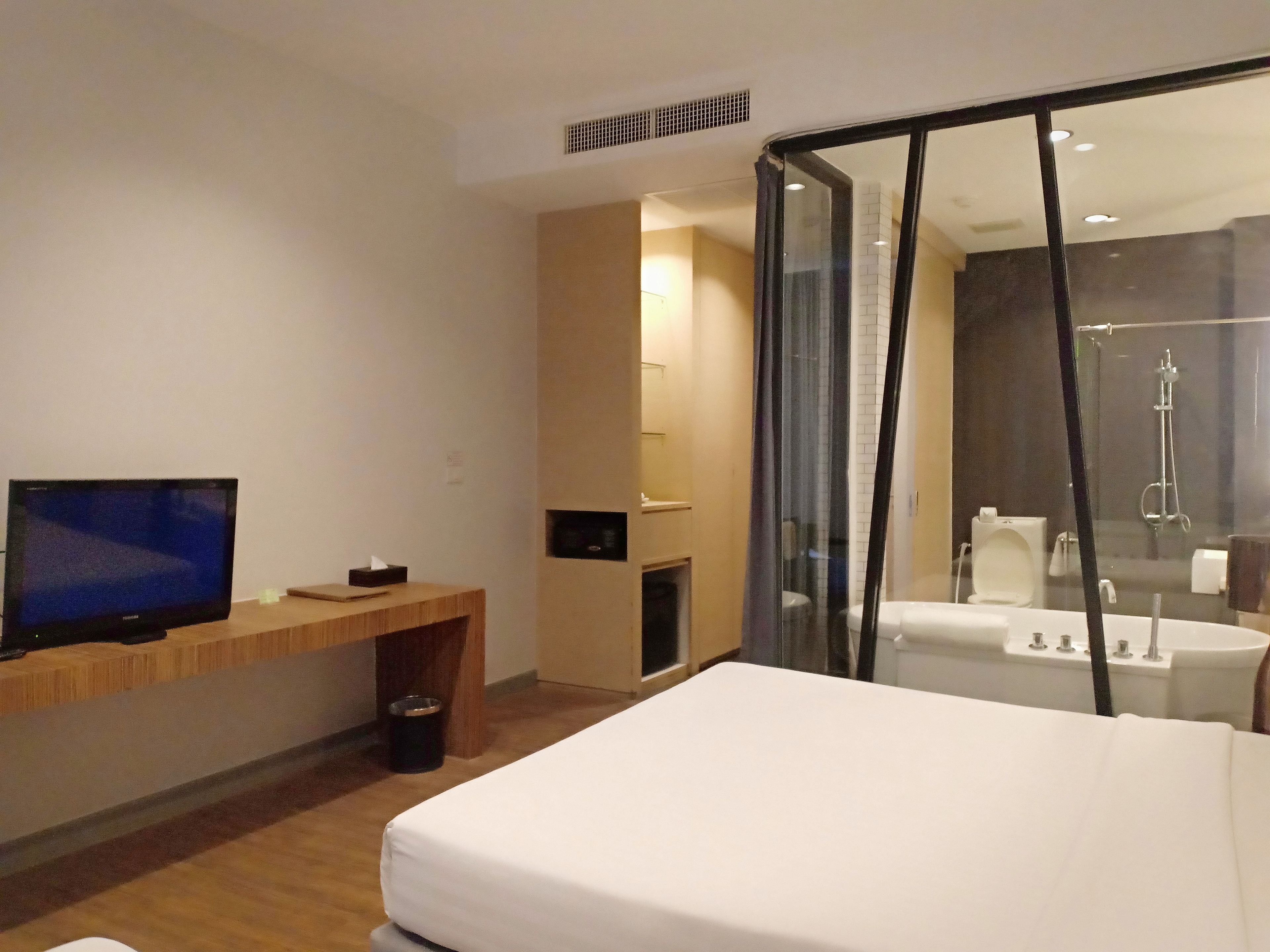 Una habitación de hotel moderna con cama, televisión, escritorio de madera, contenedor de basura y zona de ducha con paredes de vidrio.