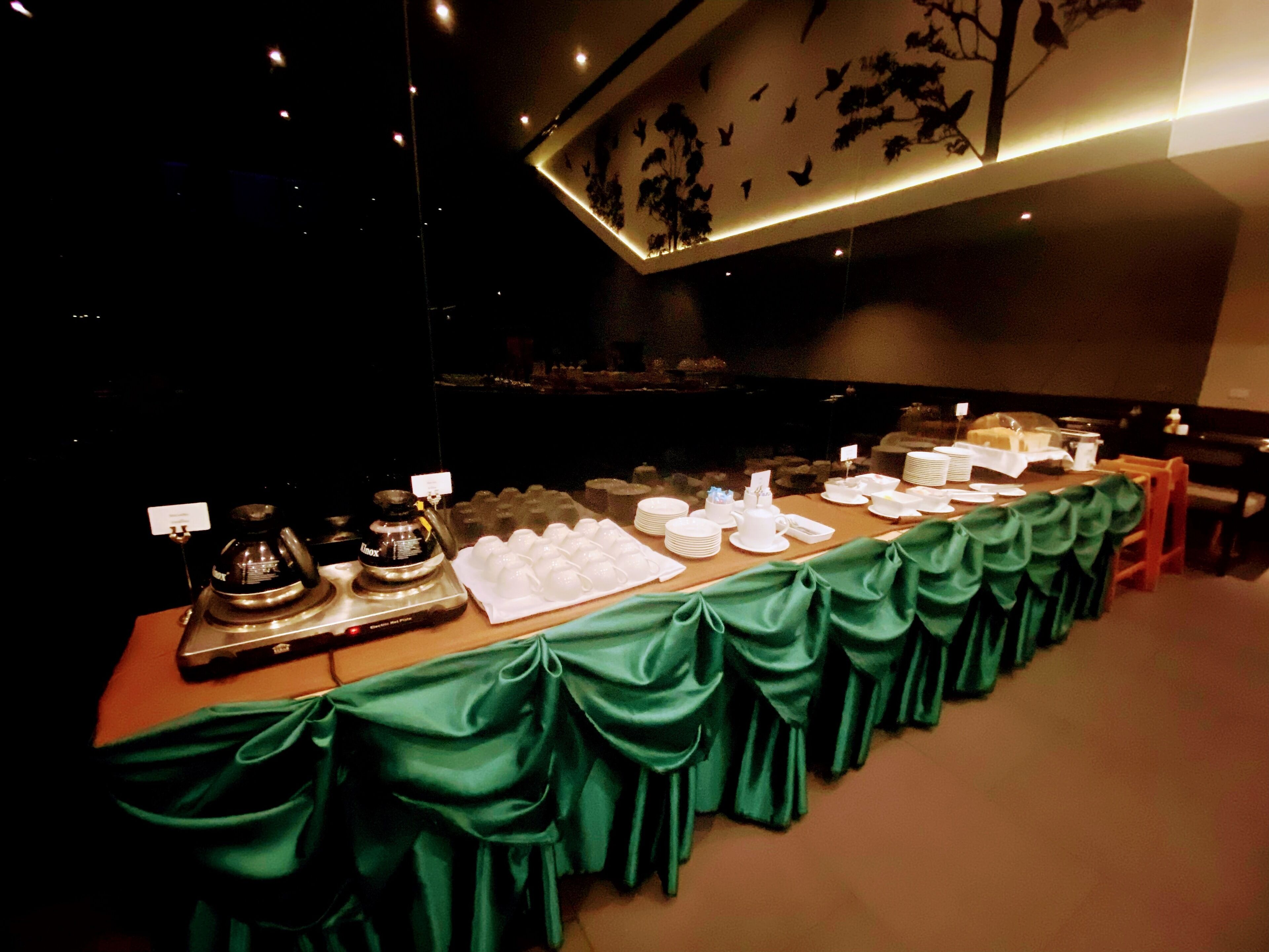 Mesa de banquete dispuesta con platos, tazas y una máquina de café, con un mantel verde drapeado y un mural de pájaros en la pared.