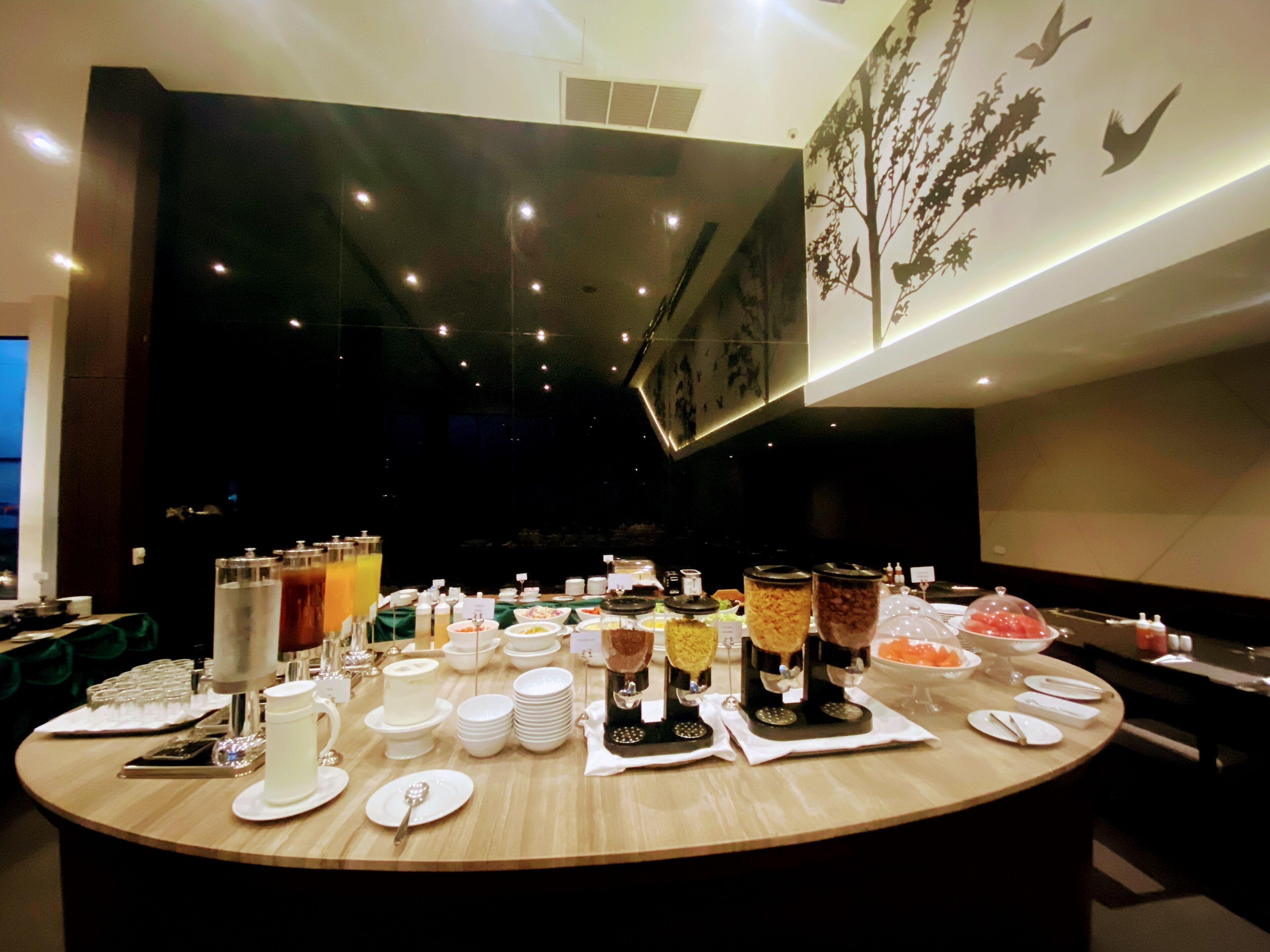A buffet setup with various food items and beverages, arranged on a round table with a backdrop of a wall with a tree mural.