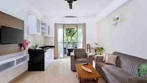 Una sala de estar moderna con un televisor de pantalla plana, un ventilador de techo, un sofá, una mesa de centro y una zona de cocina con armarios y una mesa de comedor.