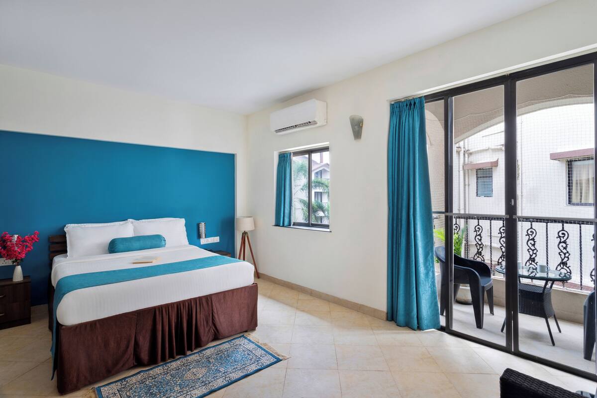 A hotel room with a bed, a small table with chairs, a balcony with a railing, and a window with curtains.