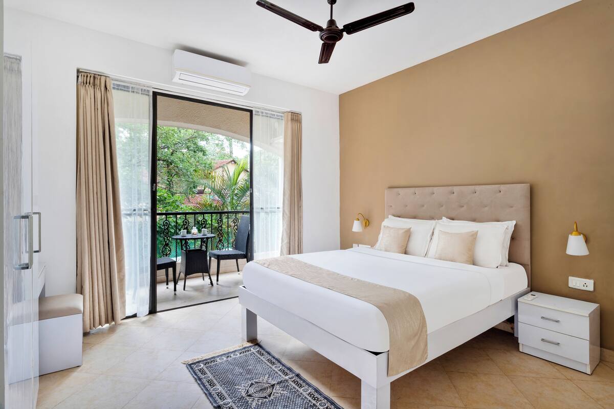 A bedroom with a bed, bedside tables, a ceiling fan, and a view of a balcony with outdoor furniture.