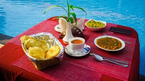 Una mesa con mantel rojo junto a una piscina, con una cesta de comida amarilla, una taza de café, un bol de curry y un teléfono inteligente.