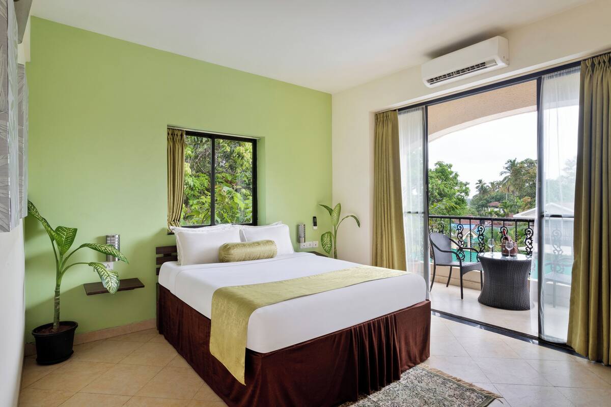 A hotel room with a bed, a small table, a potted plant, and a balcony with a view of the outdoors.