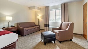 Une chambre d’hôtel avec un lit, un canapé marron, une chaise, un petit pouf, une table de chevet avec une lampe et une fenêtre avec des rideaux.