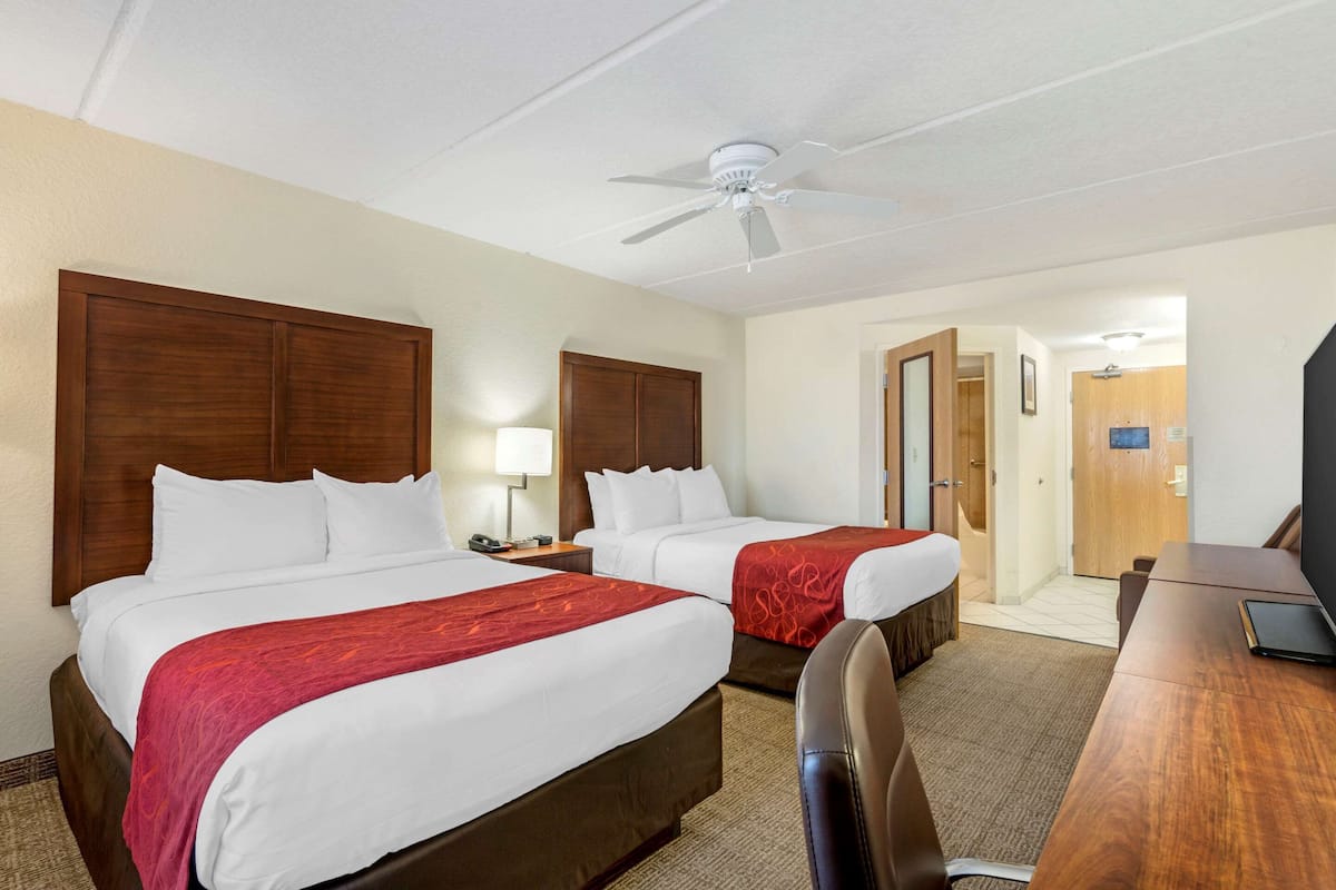 A hotel room with two beds, a desk, a chair, a ceiling fan, and a television.