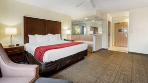 Une chambre d’hôtel avec un grand lit, un bureau avec une lampe, une chaise, un ventilateur de plafond et une salle de bain visible à travers une porte en verre.
